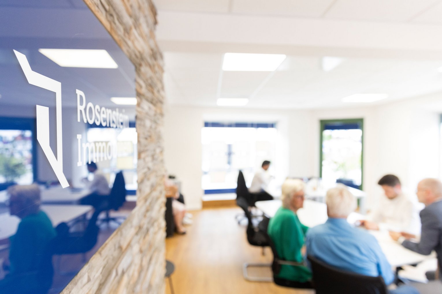 Büro von Rosenstein Immobilien Blick in das Büro von Rosenstein Immobilien, mit einem Fokus auf das Firmenlogo an der Wand und unscharfen Personen im Hintergrund bei einem Gespräch.