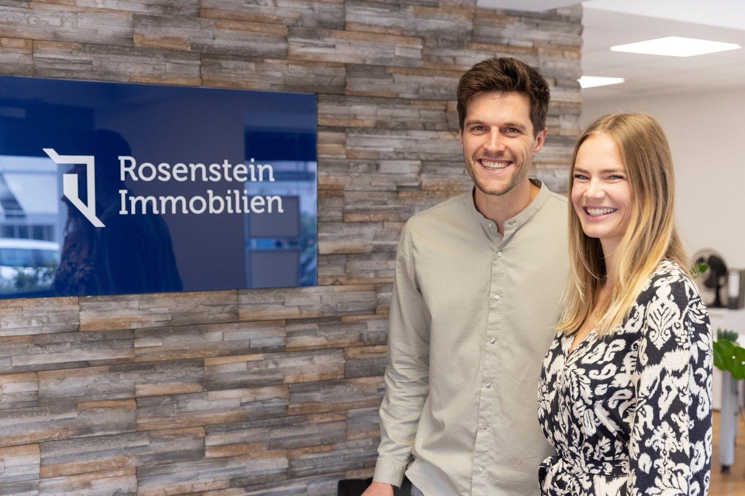 Kunden von Rosenstein Immobilien im Büro Zwei Kunden von Rosenstein Immobilien lächelnd vor dem Firmenlogo im Büro.